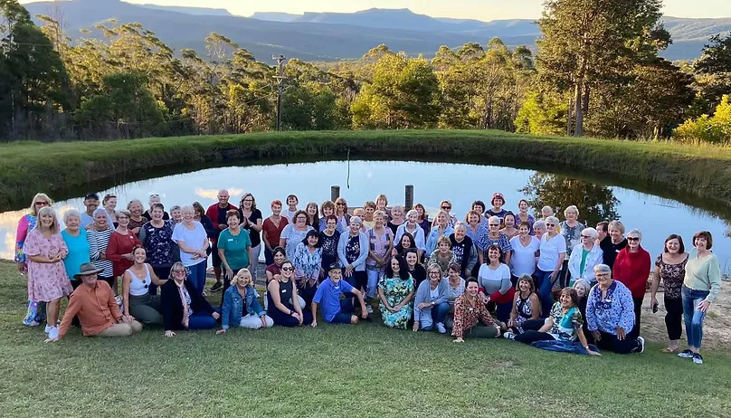 Club members posed on a grassy slope in front of a pond and mountain ridges, on a social outing.