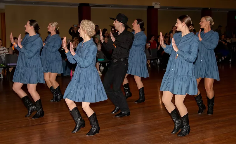 The competition team in matching denim dresses and cowboy boots, dancing in sync on a wooden floor in front of an audience.