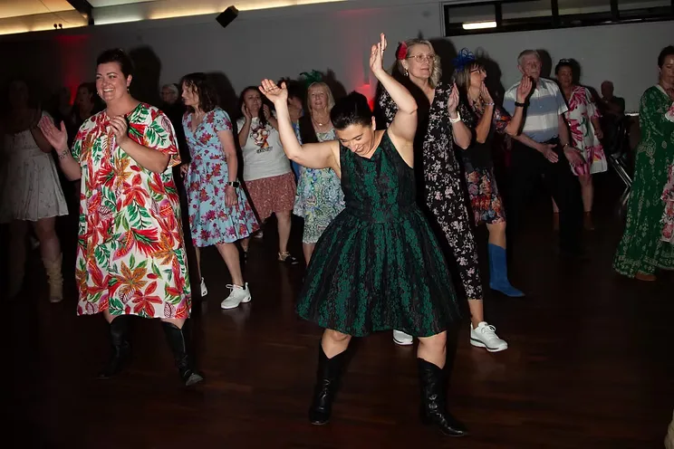 Dancers in party dresses and cowboy boots on a wooden floor, mid-routine under red hall lights.