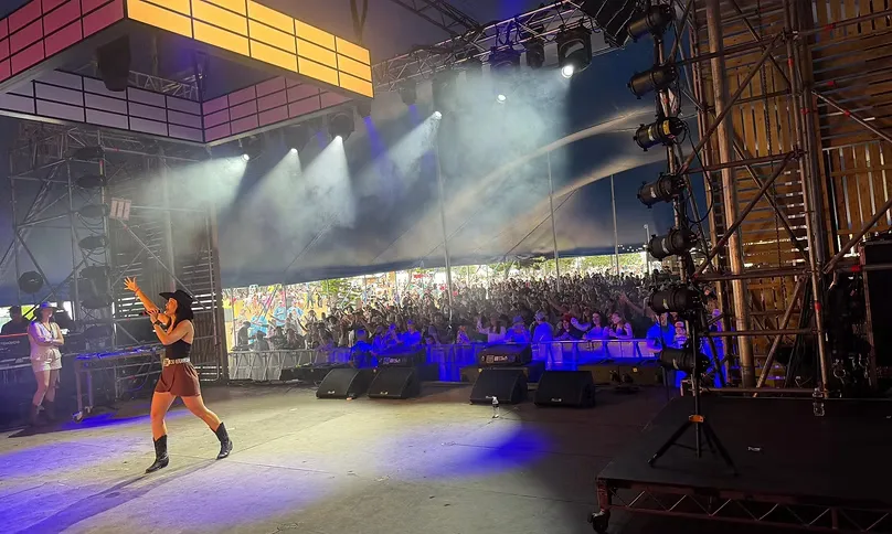 Maddison Glover on a festival stage under a marquee, hat and boots, singing into a microphone with stage lights overhead and a crowd beyond the monitors.