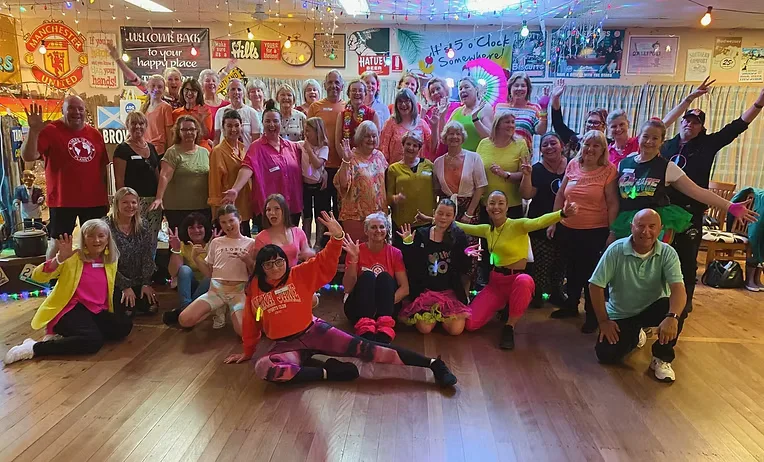 A large club group on the dance floor in neon tutus, glow sticks and colourful lights at an 80s-themed party.